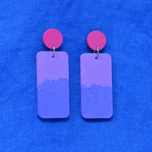 A pair of handmade drop earrings featuring rectangular purple and blue painted wooden charms with bright pink circular studs, displayed on a textured blue fabric background.