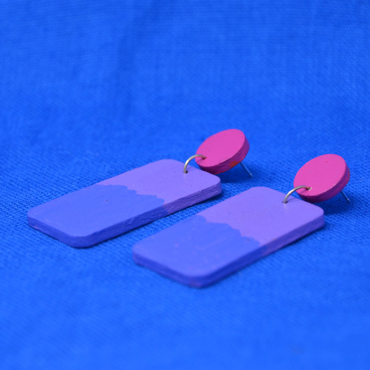 A pair of handmade wooden drop earrings featuring rectangular purple pendants with a wavy painted top border and bright pink circular studs, displayed on a textured blue background.