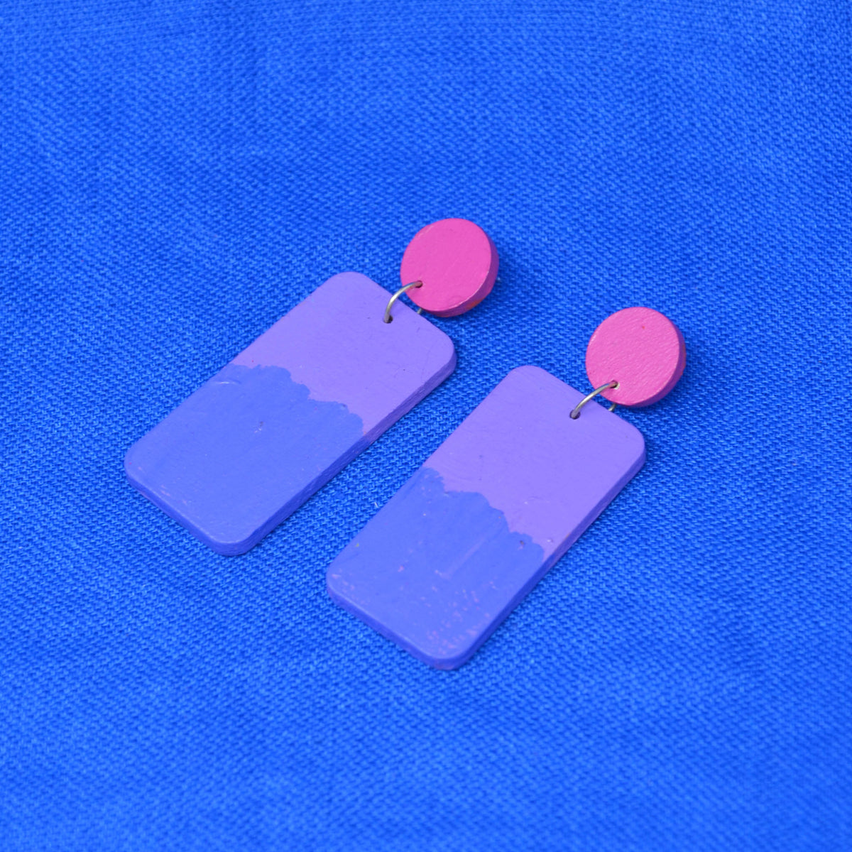 A pair of geometric drop earrings featuring rectangular purple pendants with a half-light, half-dark painted design and bright pink circular studs, displayed on a textured blue background.