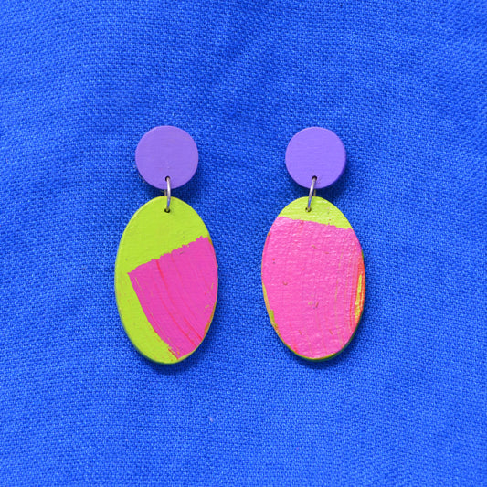 A pair of handmade drop earrings with bright neon green oval charms featuring pink and yellow painted sections, attached to purple circular studs, displayed on a textured blue background.