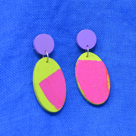 A pair of handmade drop earrings with purple circular studs and oval dangles painted in bright neon pink and lime green, displayed on a textured blue background.