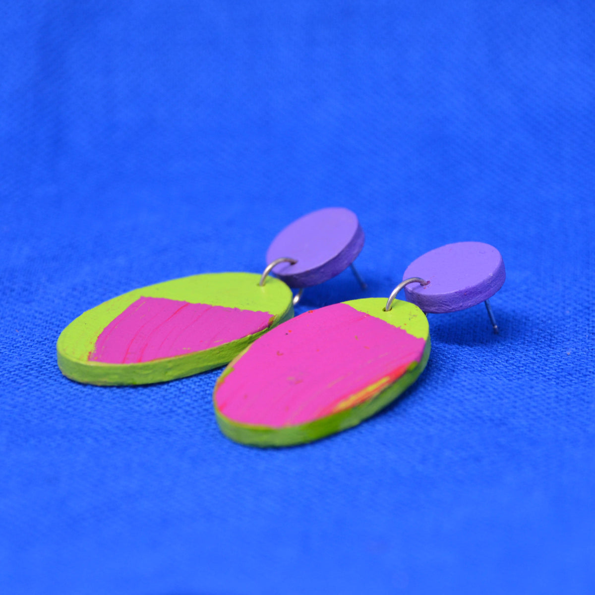 A pair of handmade wooden earrings with vibrant neon pink and lime green painted shapes, attached to small purple circular studs, displayed on a textured blue background.