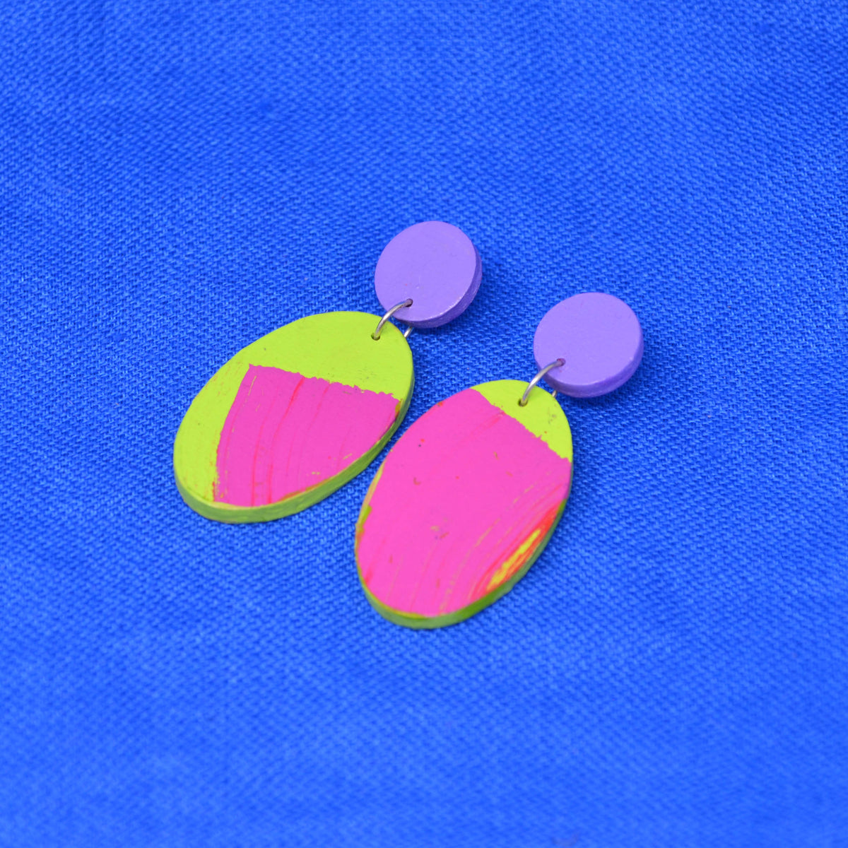A pair of drop earrings with round purple studs and oval dangles featuring a bright pink and lime green abstract color-blocked design, displayed on a textured blue background.
