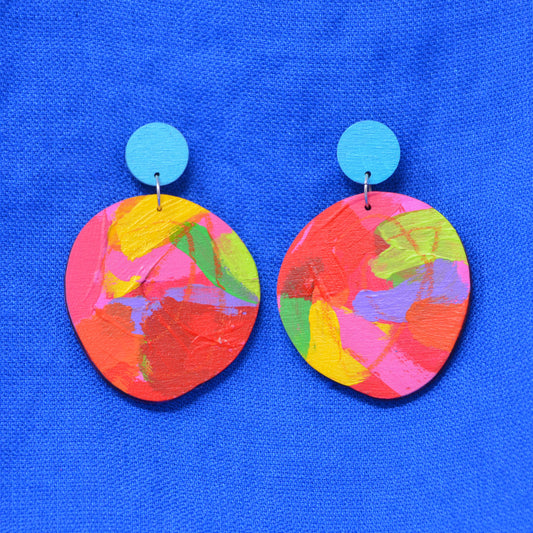 A pair of handmade earrings featuring round, abstractly painted disc charms in a vibrant multicolor mix of red, pink, yellow, green, and blue, suspended from solid turquoise blue posts, displayed on a textured blue background.