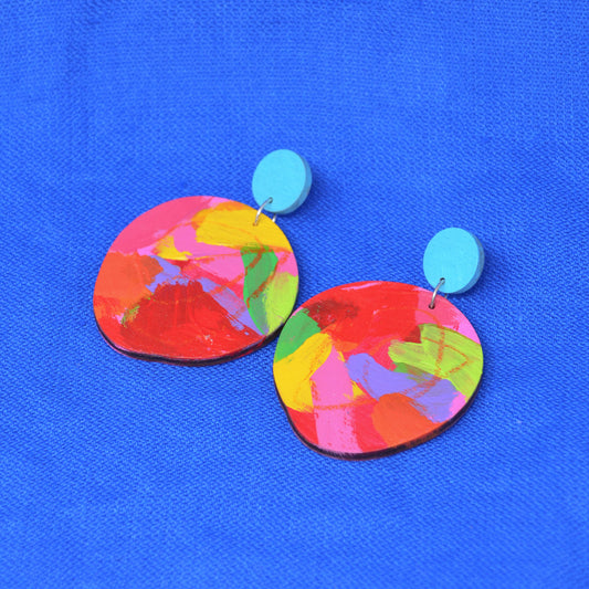 A pair of colorful abstract dangle earrings with round turquoise blue studs and irregular oval drops featuring a vibrant multicolor paint pattern, displayed on a textured blue background.