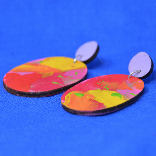 A pair of handmade oval drop earrings featuring a colorful abstract paint design in red, yellow, orange, and pink, with a solid lavender-purple circular back and silver-colored studs, displayed on a bright blue textured background.