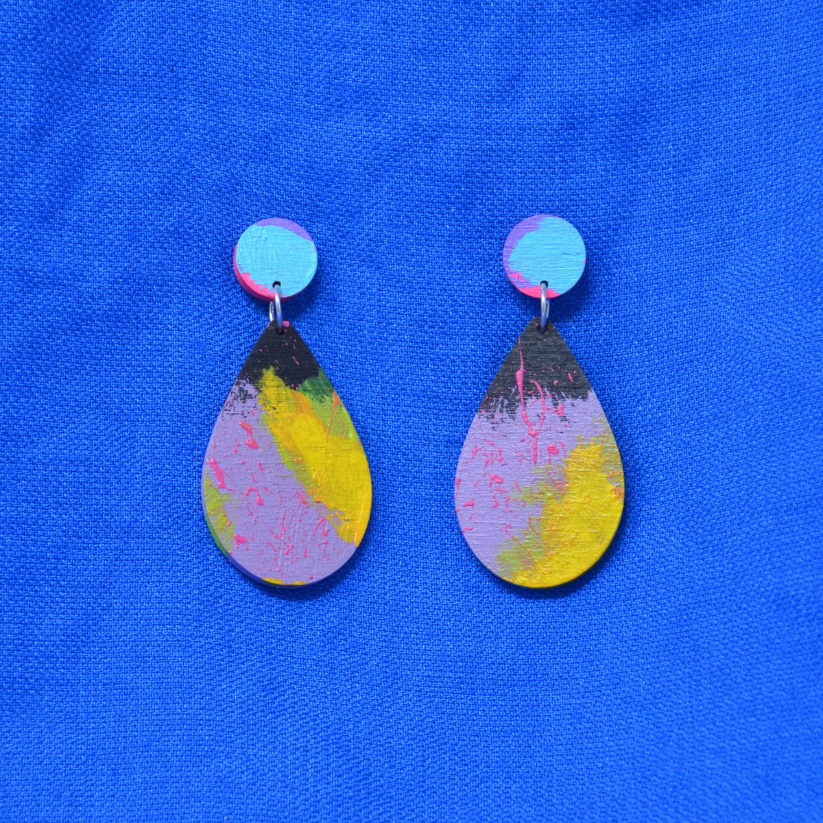 A pair of handmade teardrop dangle earrings with a colorful abstract paint design in yellow, pink, purple, and black, featuring light blue circular studs, displayed on a textured blue background.