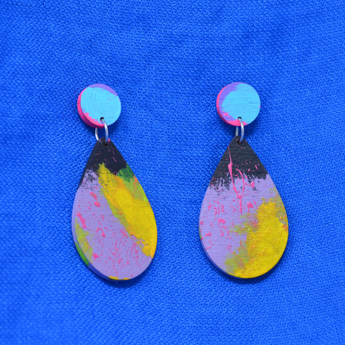A pair of handmade teardrop dangle earrings with a colorful abstract paint design in pink, yellow, purple, and black, hanging against a bright blue textured fabric background.