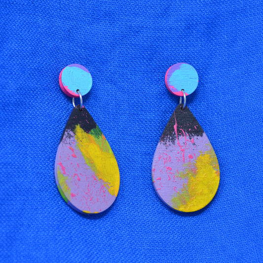 A pair of handmade teardrop dangle earrings with a colorful abstract paint design in pink, yellow, purple, and black, hanging against a bright blue textured fabric background.