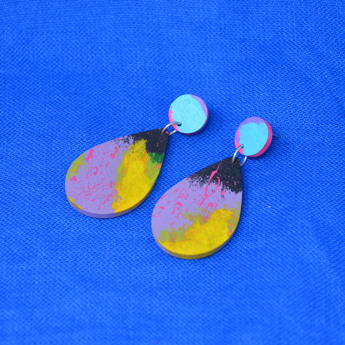 A pair of handmade teardrop-shaped drop earrings with a colorful abstract paint design in yellow, pink, purple, and black, hanging from light blue circular studs on a textured blue background.