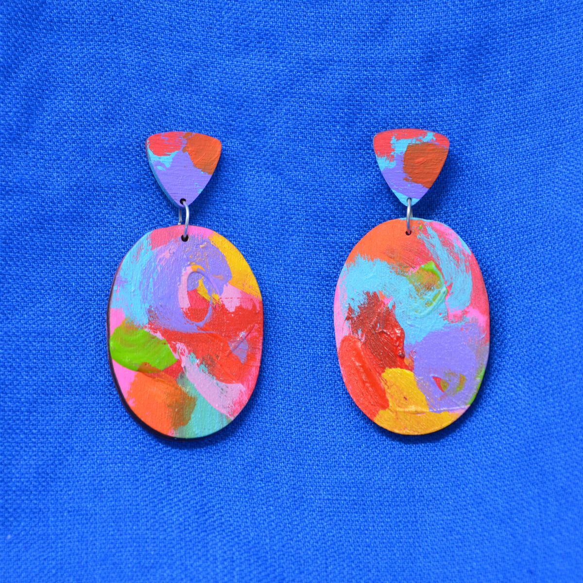 A pair of handmade drop earrings with abstract multicolor painted oval pendants and matching painted triangular studs, displayed on a textured blue fabric background.