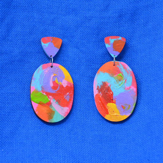 A pair of handmade drop earrings with abstract multicolor painted oval pendants and matching painted triangular studs, displayed on a textured blue fabric background.