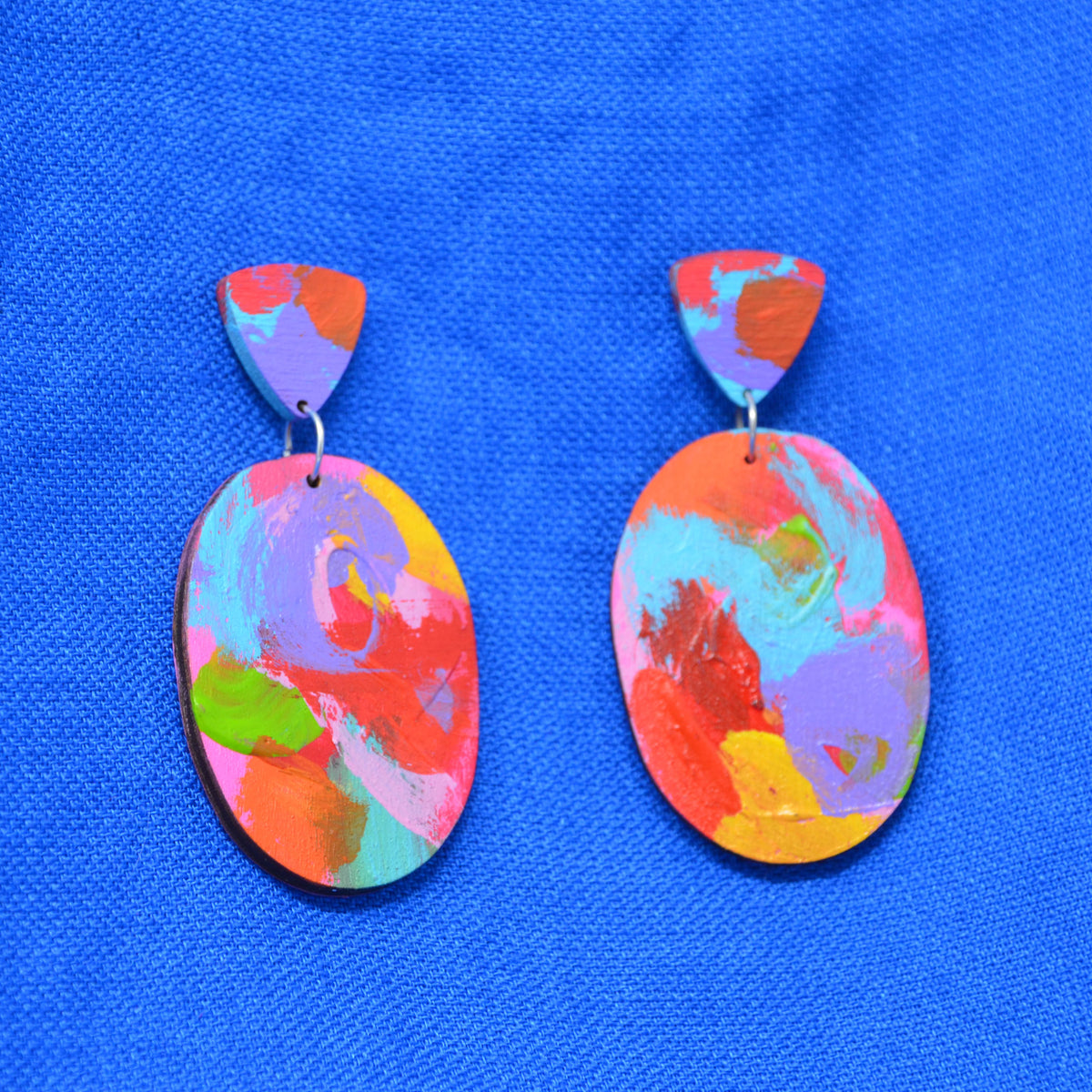 A pair of colorful abstract painted earrings with oval drop pendants and triangular top pieces, featuring swirls of pink, orange, purple, blue, yellow, and green on a bright blue textured background.