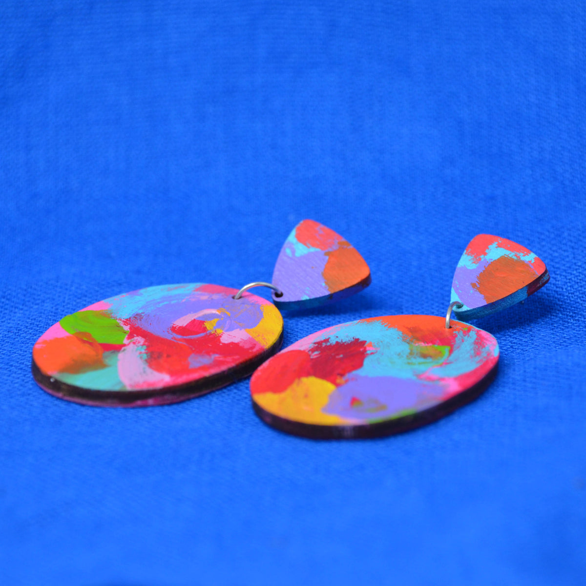 A pair of colorful abstract dangle earrings with oval and triangular shapes, featuring a vibrant paint-splatter pattern in red, orange, yellow, green, blue, and purple, displayed on a textured blue background.