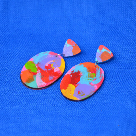 A pair of colorful abstract painted earrings with irregular oval drops and small triangular studs, shown on a bright blue textured background.