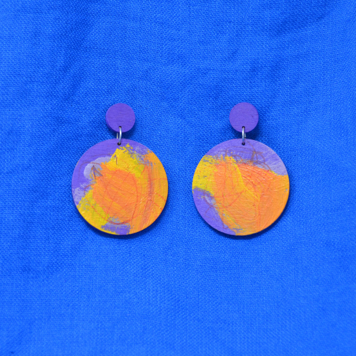 A pair of handmade drop earrings with purple circular studs and round dangles featuring an abstract marbled pattern of purple, orange, and yellow, displayed on a textured blue fabric background.