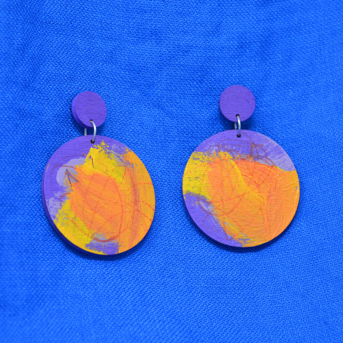 A pair of handmade wooden dangle earrings with purple circular studs and larger round wooden discs featuring an abstract orange and yellow painted pattern, displayed on a textured blue background.