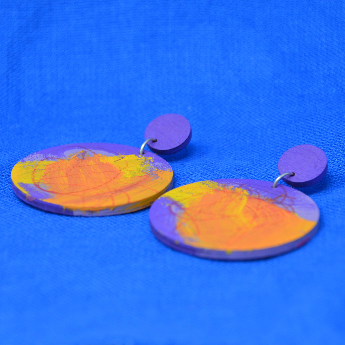 A pair of handmade disc earrings with a purple and orange marbled pattern, featuring a small round purple disc stud, displayed on a textured blue background.
