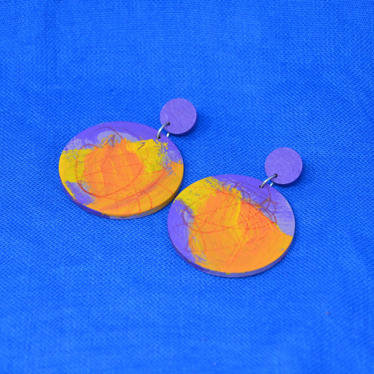 A pair of handmade round drop earrings featuring a purple and orange marbled pattern with fine gold vein details, displayed on a textured blue background.