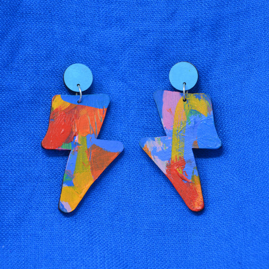 A pair of lightning bolt-shaped drop earrings with a colorful abstract paint design and light blue circular studs, displayed on a textured blue background.