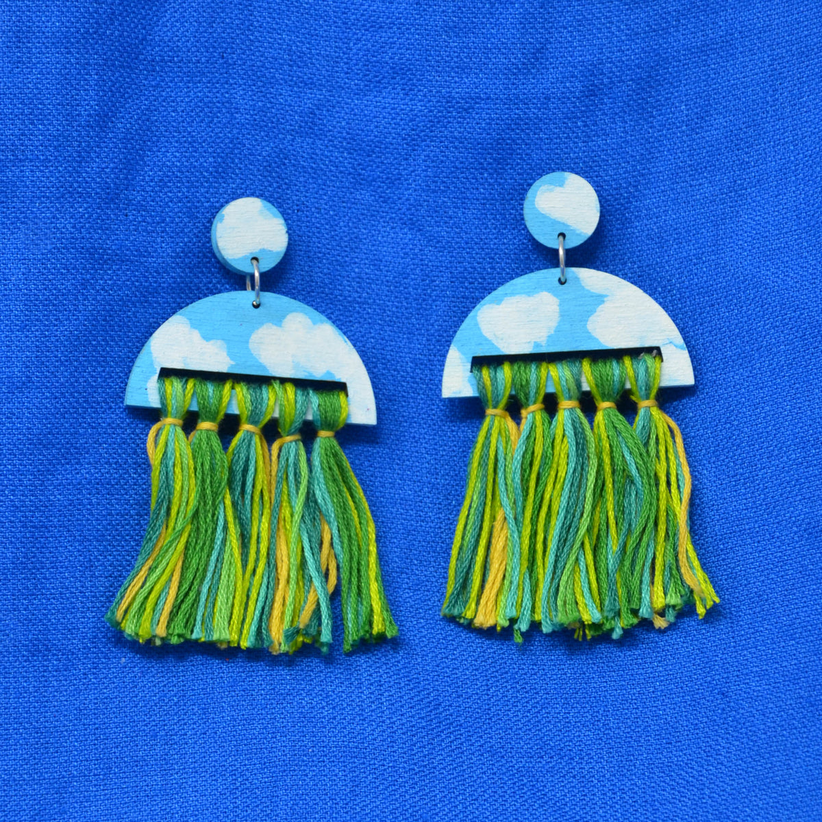A pair of handmade dangle earrings featuring sky-blue and white cloud-patterned semicircles with green and yellow tassels, displayed on a textured blue fabric background.