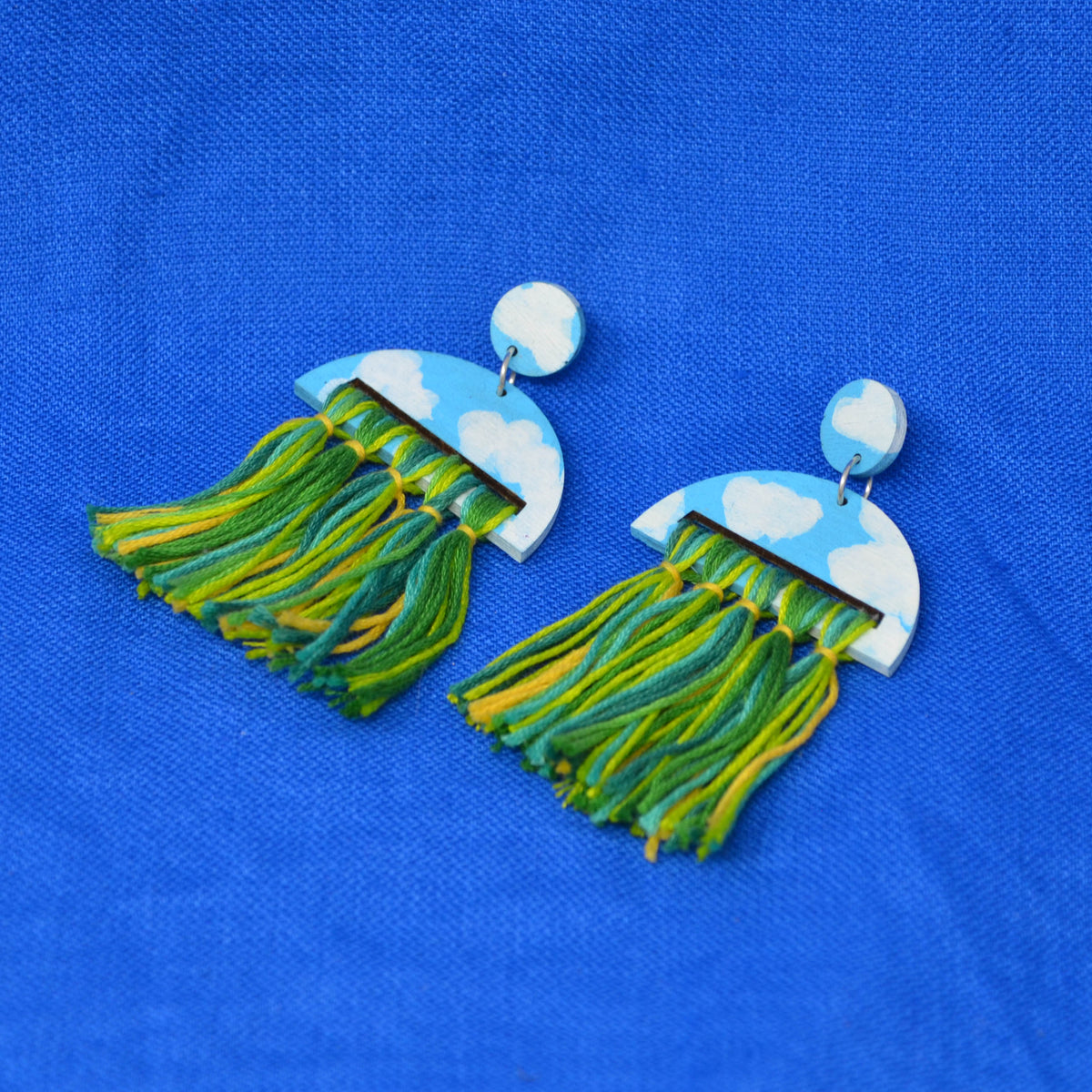 A pair of jellyfish-shaped drop earrings with blue and white cloud-patterned tops and long green and yellow fringe tassels, displayed on a textured blue fabric background.