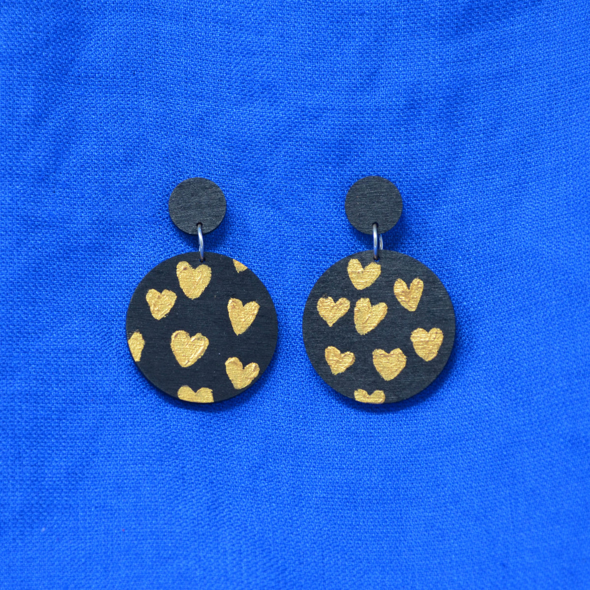 A pair of handmade drop earrings featuring round black wooden discs decorated with gold-painted hearts, displayed on a vibrant blue textured fabric background.