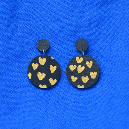 A pair of handmade drop earrings featuring round black wooden discs decorated with gold-painted hearts, displayed on a vibrant blue textured fabric background.