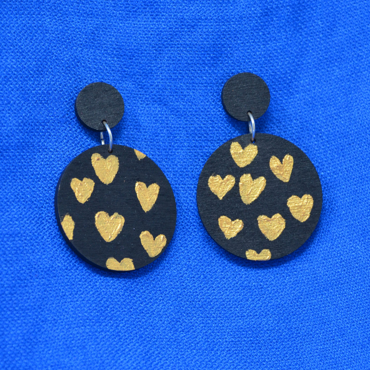 A pair of handmade drop earrings featuring round black wooden discs decorated with hand-painted gold hearts, displayed on a vibrant blue textured fabric background.
