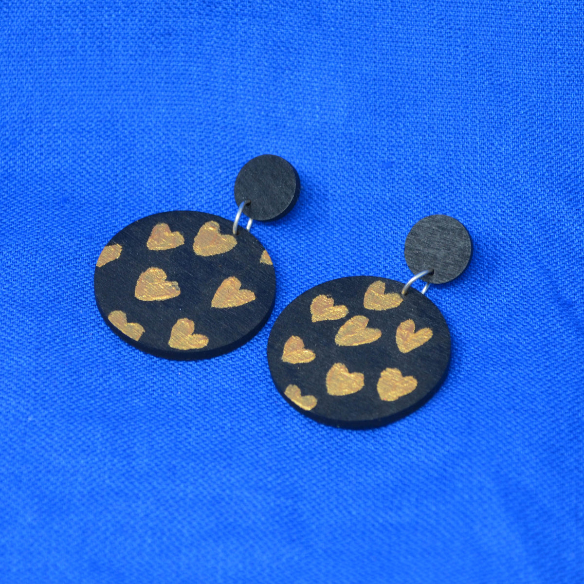 A pair of handmade drop earrings featuring round black wooden discs decorated with gold hearts, suspended from small black metal studs, displayed on a textured blue background.