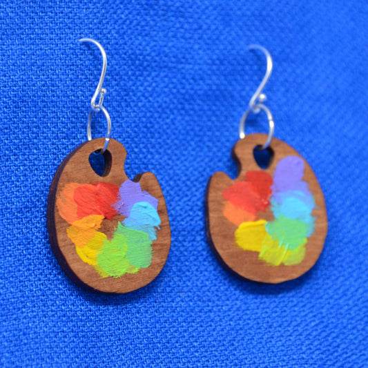 A pair of wooden paint palette earrings with colorful hand-painted paint strokes, hanging from silver fishhook wires against a textured blue background.