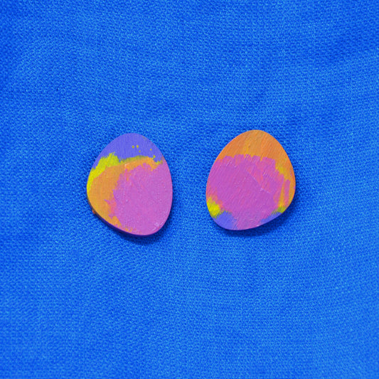 Two small oval painted wooden earring with pink, purple, orange, and yellow colors, placed side by side on a bright blue textured fabric background.