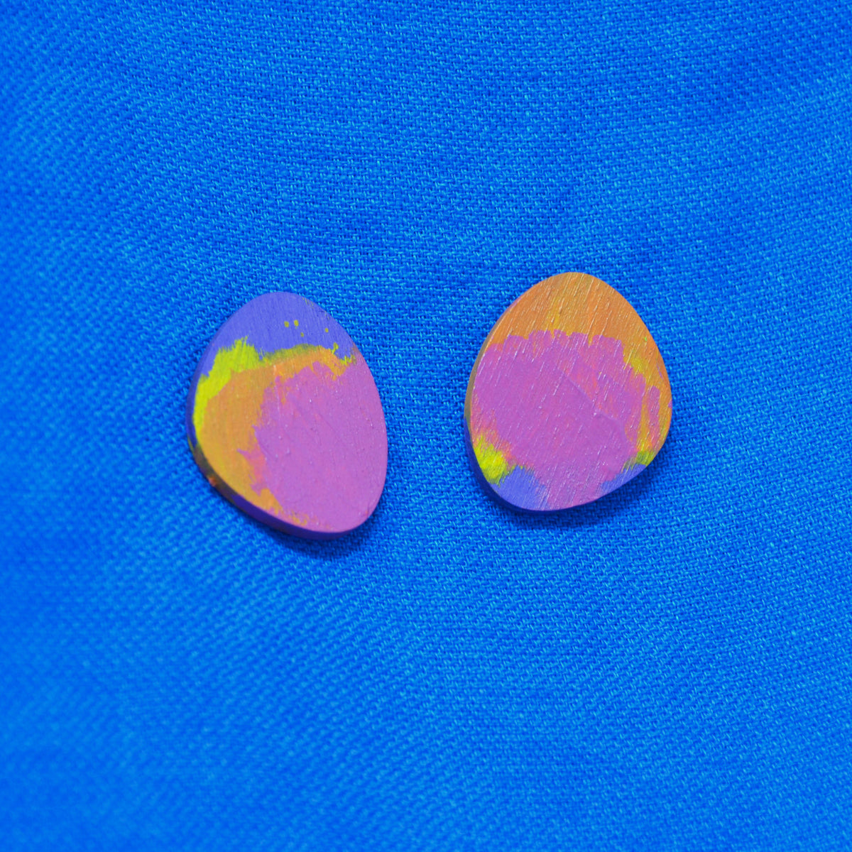Two  teardrop-shaped wooden earrings with a marbled pink, purple, orange, and yellow pattern, displayed on a bright blue textured fabric background.