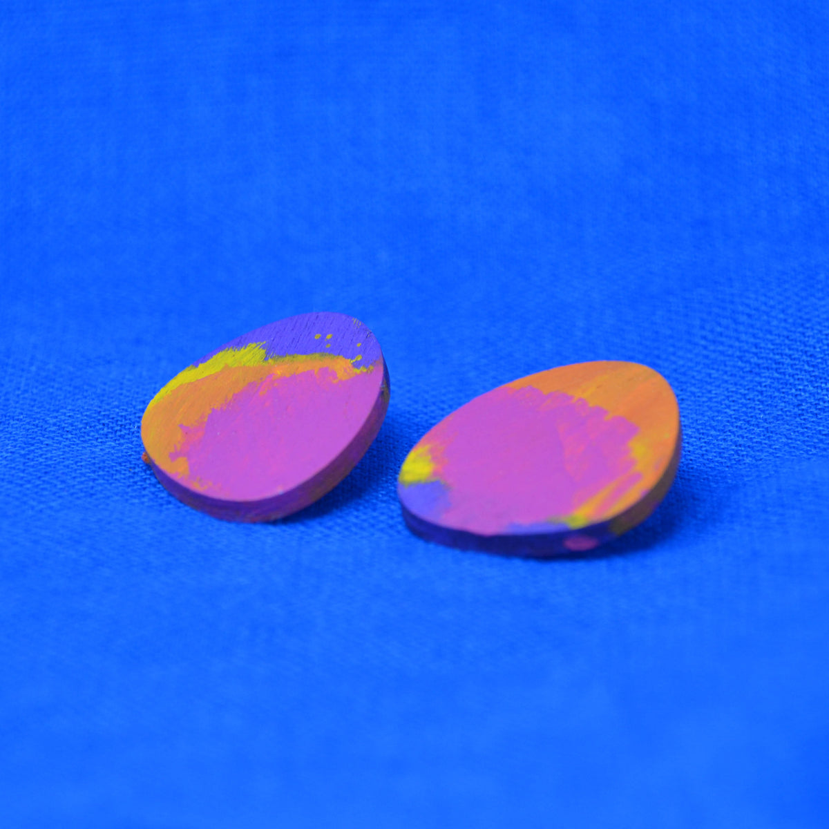 Two small teardrop-shaped wooden beads with a marbled pattern of purple, pink, orange, and yellow, displayed on a textured blue background.