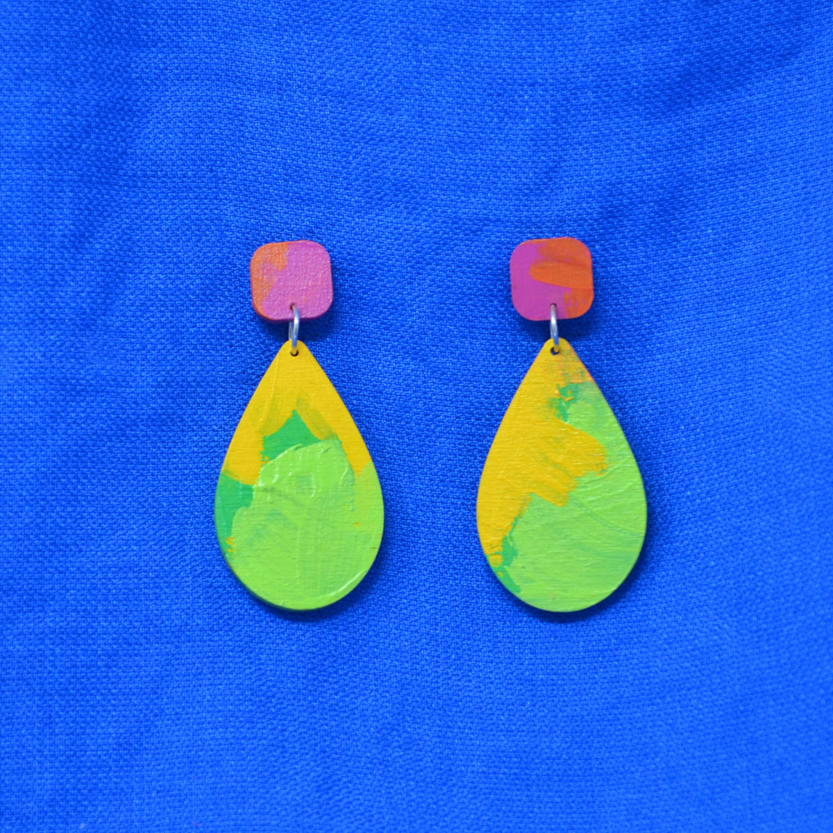 A pair of handmade teardrop earrings with bright yellow and green painted drop shapes and pink square posts, displayed on a textured blue background.
