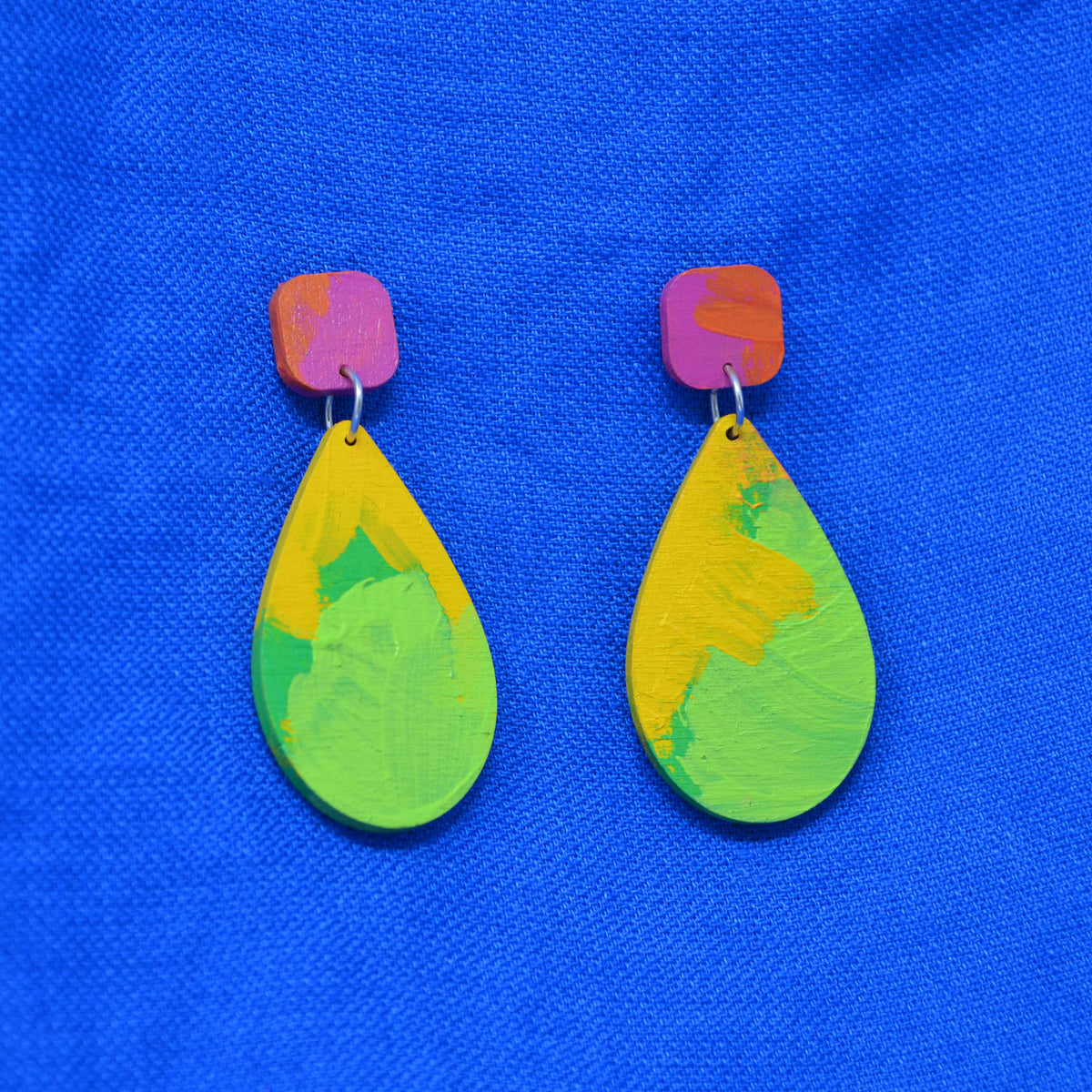 A pair of handmade teardrop earrings with vibrant, multicolored hand-painted wooden drops and pinkish-orange painted posts, displayed on a textured blue fabric background.