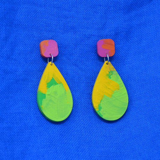 A pair of handmade teardrop earrings with vibrant, multicolored hand-painted wooden drops and pinkish-orange painted posts, displayed on a textured blue fabric background.