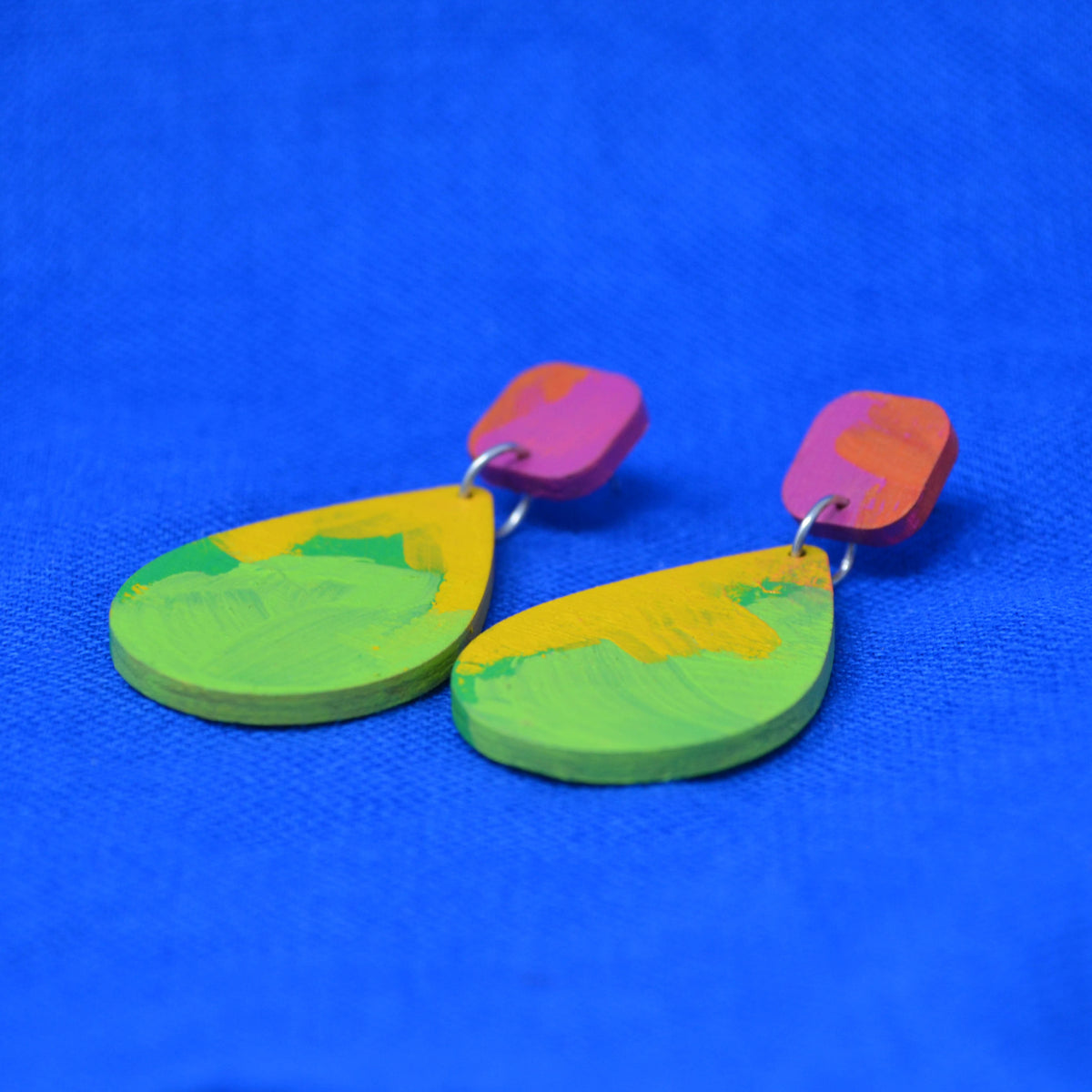A pair of handmade wooden dangle earrings with colorful, textured paint designs in green, yellow, pink, and orange, displayed on a vibrant blue textured background.