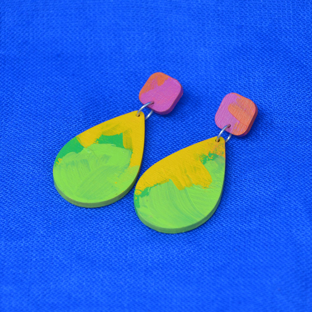 A pair of handmade teardrop dangle earrings with a colorful, textured paint-stroke design in yellow, green, and pink, displayed on a bright blue textured fabric background.