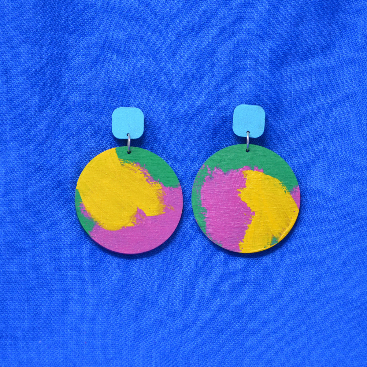 A pair of wooden drop earrings with colorful circles, with hand-painted paint strokes, hanging from a rounded light blue square stud, against a textured blue background.
