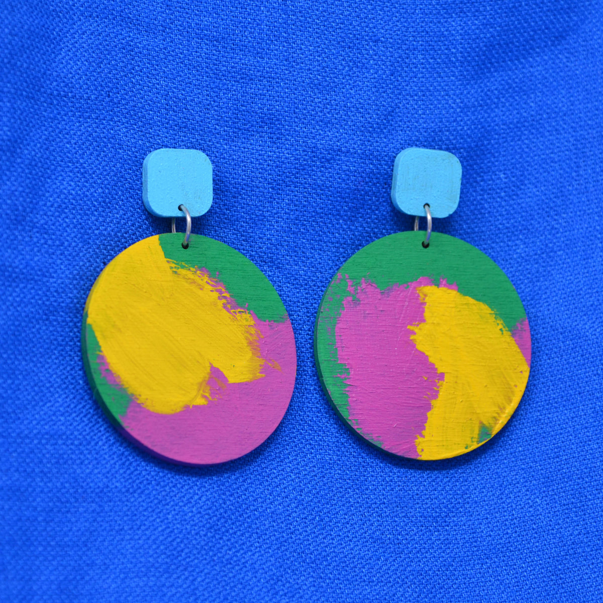 A pair of wooden drop earrings with colorful circles, with hand-painted paint strokes, hanging from a rounded light blue square stud, against a textured blue background.