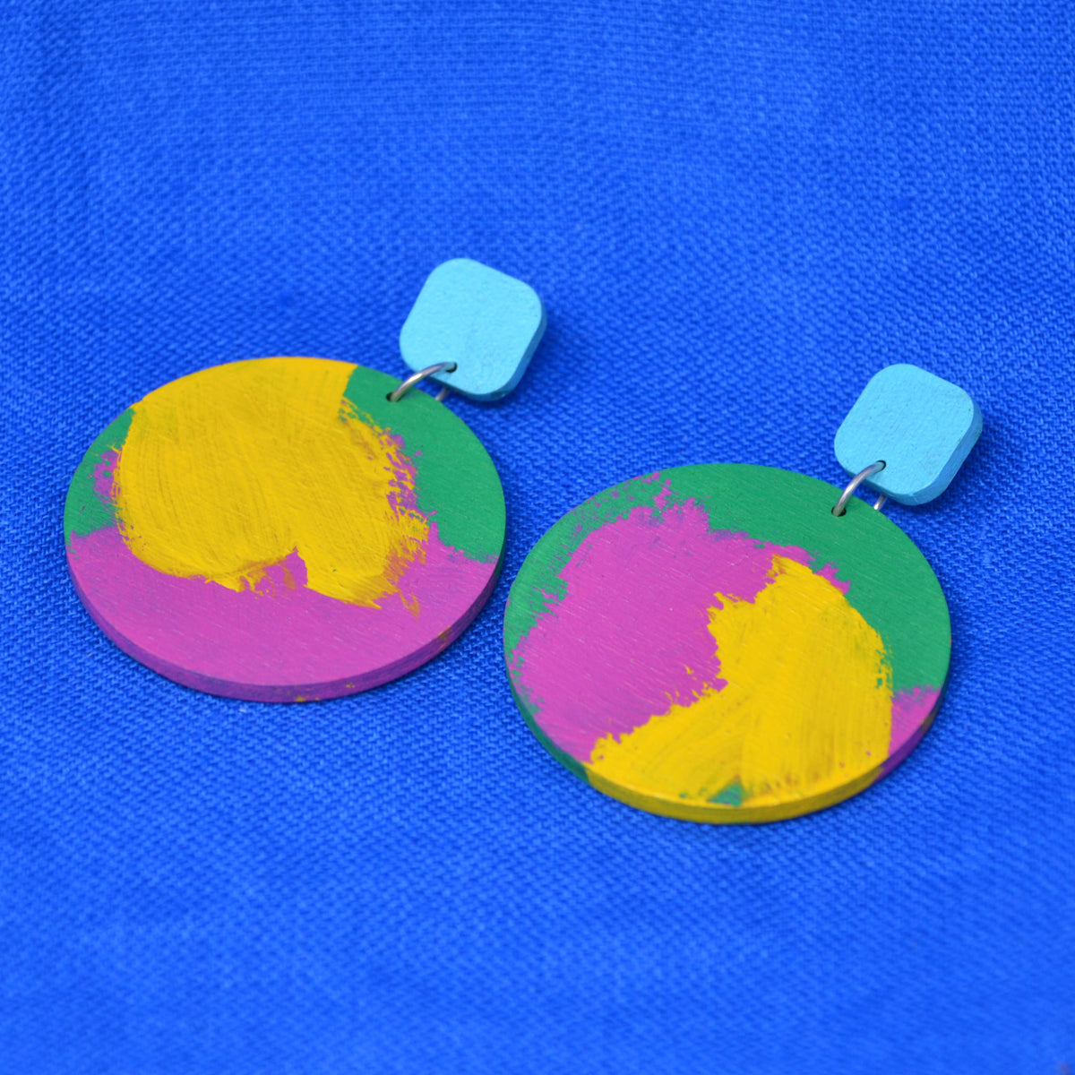 A pair of wooden drop earrings with colorful circles, with hand-painted paint strokes, hanging from a rounded light blue square stud, against a textured blue background.