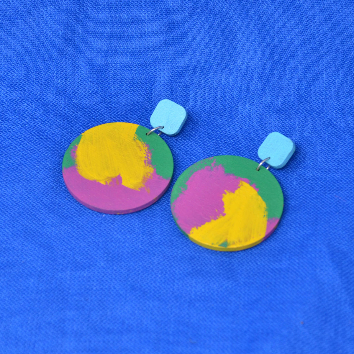 A pair of wooden drop earrings with colorful circles, with hand-painted paint strokes, hanging from a rounded light blue square stud, against a textured blue background.