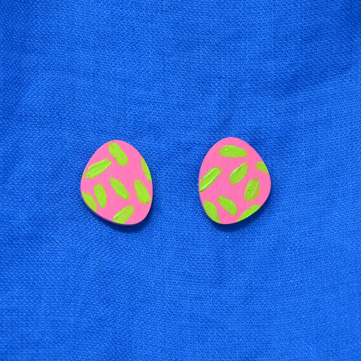 A pair of pink and green stud earrings, displayed on a textured blue fabric background.