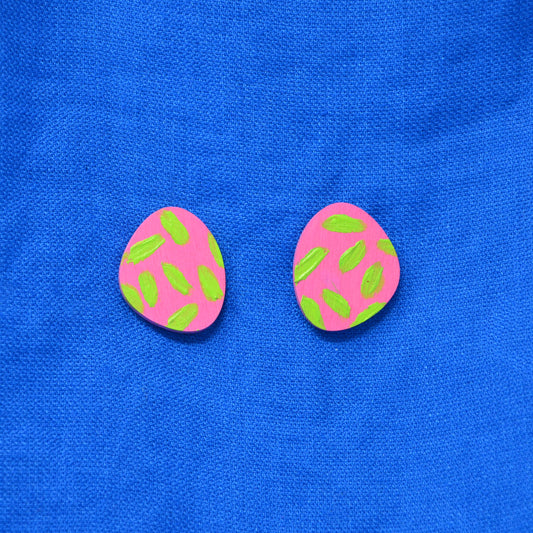 A pair of pink and green stud earrings, displayed on a textured blue fabric background.