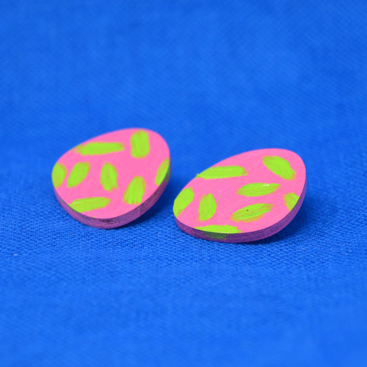 A pair of small, oval-shaped wooden earring studs with a bright pink background and lime green detail, displayed on a textured blue fabric background.