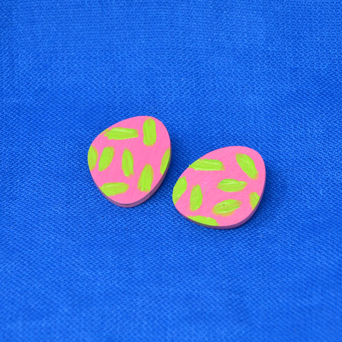 Pair of pink and green painted stud earrings, displayed on a textured blue background.