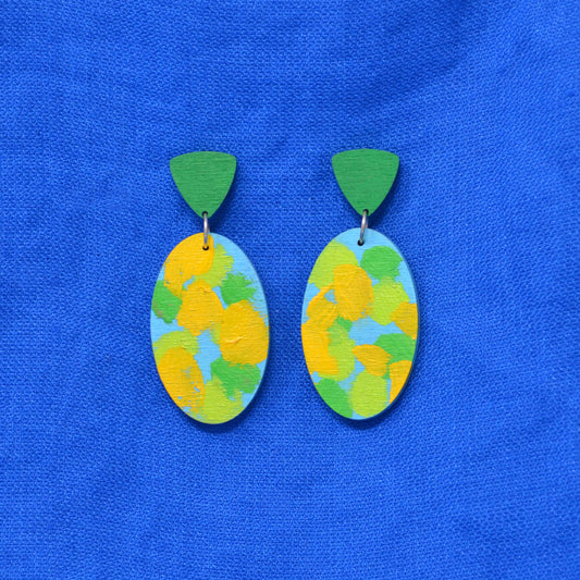 A pair of handmade wooden dangle earrings with green triangle-shaped tops and oval drop charms painted with yellow, green, and light blue brushstrokes, displayed on a textured blue fabric background.