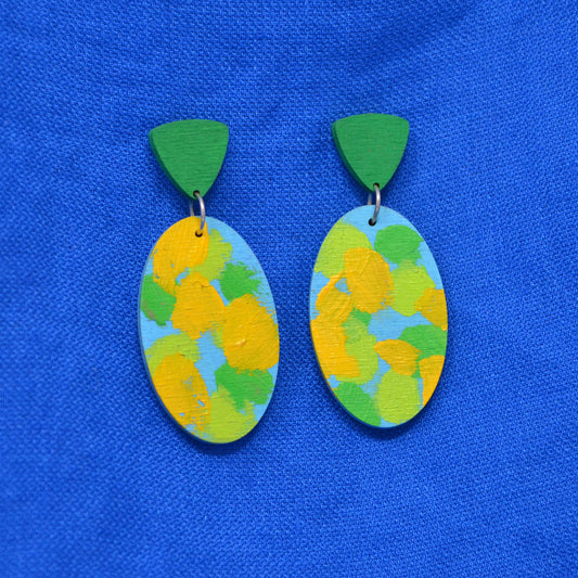 A pair of handmade wooden drop earrings with green triangular studs and oval dangles painted with yellow, green, and light blue brushstrokes, displayed on a textured blue fabric background.