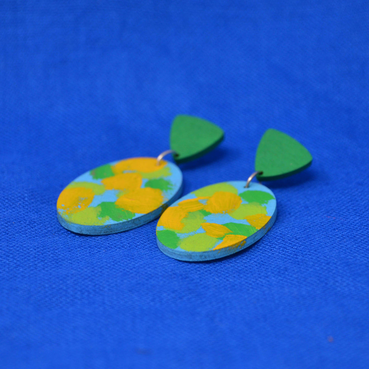 A pair of handmade wooden dangle earrings with green triangular posts and oval drops featuring a blue, green, and yellow abstract paint pattern, displayed on a textured blue background.